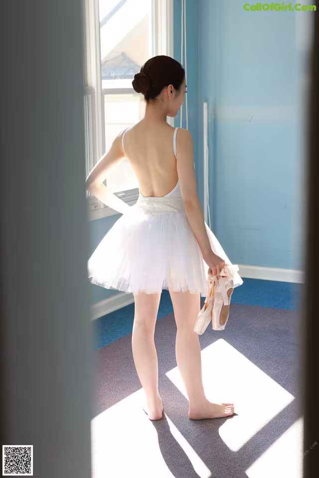 A young ballerina standing in front of a window holding a pair of ballet shoes.