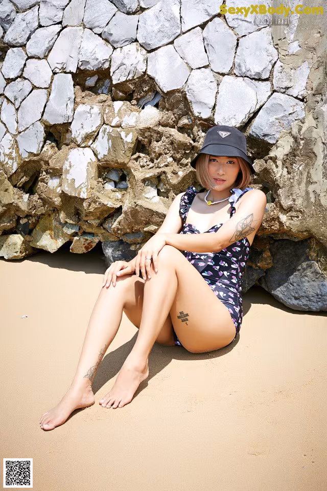 A woman sitting on the sand in front of a stone wall.